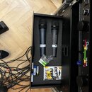 Otevřený černý kufřík s pěnovou vložkou, ve kterém jsou umístěny dva černé bezdrátové mikrofony. Na jednom z nich je vidět bílý nápis „SENNHEISER“ a…