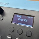 Detailní záběr na horní část trigger modulu v modré a černé barvě. Na modrém panelu je nápis TRIGGER MODULE TM-6 PRO. Digitální displej zobrazuje tex…
