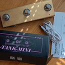 Fotografie zobrazuje zlatý efekt pedál s třemi stoupacími spínači, černou krabici a bílý uživatelský manuál s přiloženým USB kabelem. Na krabici je f…