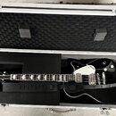 Otevřený hliníkový kufr s černou pěnovou výplní, ve kterém leží černá elektrická kytara a černý popruh. Na hlavě kytary je nápis GRETSCH. Tělo kytary…