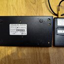 Fotografie zachycuje zadní stranu černého přijímače bezdrátového mikrofonu a přilehlý napájecí adaptér. Na přijímači je bílý štítek s nápisy WIRELESS…