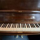 Detail klaviatury a horní části tmavě hnědého dřevěného pianina. Uprostřed nad klávesami je vyryt nápis PETROF. Bílé klávesy vykazují mírné zežlutnut…
