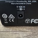 Detailní pohled na zadní černou plastovou stranu zařízení s napájecím vstupem. Je zde bílý nápis Designed in Woodinville, WA, USA. Manufactured in Ch…