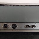 Zadní strana šedého elektronického zařízení s kovovým panelem v dolní části. Na panelu je napájecí konektor, přepínač s nápisy 110, 130, 220, 240 a o…