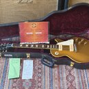Zlatá elektrická kytara typu hollow body v černém tvrdém kufru s červeným vnitřkem. Na kytaru jsou vidět dva humbuckery a čtyři ovládací knoflíky. V…