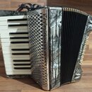 Stříbrný akordeon s bílými a černými klávesami. Na bočnici je svisle napsán nápis HOHNER a na pravém okrahu nápis STUDENT III. Povrch nástroje vykazu…