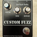 Černý efekt pedál s nápisem CUSTOM FUZZ a značkou DevTech Audio. Obsahuje tři stříbrné oválné knoflíky s popisky VOLUME, TONE a SUSTAIN. V horní část…