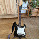 Černá elektrická kytara typu Stratocaster s bílým pickguardem a hnedým hmatníkem. Na hlavě je vidět nápis "Fender Stratocaster". Nástroj stojí na čer…