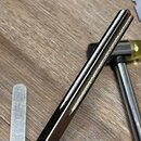 Detailní záběr na kovový nástroj s drážkovanou pracovní plochou a černou rukojetí. Vedle nástroje je vidět část kladiva s žlutou hlavou a kovový piln…