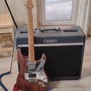 Elektrická kytara s hnědým dřevěným povrchem a bílým pickguardem stojí před černým kombem. Na hlavě kytary je nápis STRATOCASTER. Kombo má na přední…