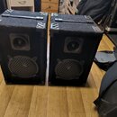 Dva černé reproduktorové boxy s texturovaným povrchem a kovovými hranami. Na přední mřížce každého reproduktoru je viditelné logo JBL. Povrch boxů vy…