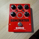 Červený efekt pedál s nápisy DOMINATOR a OHKO HANDBUILT IN GERMANY. Obsahuje pět černých otočných ovladačů s popisky BASS, MIDS, TREBLE, GRAIN a LEVE…