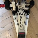 Fotografie zachycuje detail bicí pedál v bílé barvě s kovovými prvky. Na stupni je černý nápis „dw 9000“ a pod ním text „The Drummer's Choice.®“. Ped…