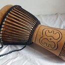 Dřevěný buben typu djembe s přirozenou barvou dřeva a černým provázkem. Na těle nástroje je vyrytý ornament ve tvaru stylizovaného symbolu a geometri…