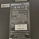 Detailní pohled na zadní část černého digitálního pianina s připojeným pedalem. Na přístroji jsou viditelné štítky se značkou Roland a modelem F701.…