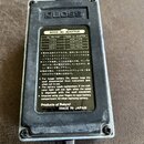 Fotografie zachycuje zadní stranu stříbrného efekt pedálu s kovovým šasi. Na černé štítku jsou nápisy BOSS AC ADAPTOR, tabulka s napájecími adaptéry…