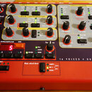 Červený syntetizátor s množstvstvím ovládacích knoflíků, přepínačů a nápisy. Na panelu jsou vidět názvy sekcí jako OSCILLATORS, AMPLIFIER, FILTER a L…