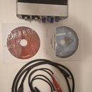 Černý audio rozhraní s hliníkovým předním panelem a několika konektory. Vedle něj jsou dva kompaktní disky, z nichž jeden nese nápisy FIREBOX a PreSo…