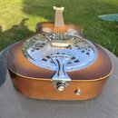 Akustická kytara s hnědým přechodovým lakem (sunburst) a kovovou krycí deskou s otvory. Na chromovaném krytu je vidět nápis "Resonator". Povrch nástr…