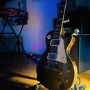 Černá elektrická kytara typu Les Paul s bílým pickguardem a dvěma humbuckery, umístěná na stojanu. V pozadí je vidět černý kombozesilovač se zlatým n…