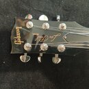 Detail hlavy černé elektrické kytary s nápisy Gibson a MODEL Les Paul v gold barvě. Na hlave jsou vidět stříbrné ladicí kolíky a struny vedené přes m…