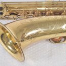 Detail zlatého saxofonu s vyrytým ornamentem na zvonu. Na povrchu je čitelný nápis YASAGUSA JAPAN. Nástroj vykazuje drobné škrábance a známky opotřeb…