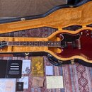 V otevřeném černém tvrdém kufru s žlutým vnitřkem leží tmavě červená elektrická kytara typu semi-hollow s černým pickguardem. Na hlavě hmatníku je bí…