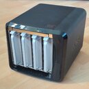 Černý, kostkovitý elektronický přístroj s nápisem Zyxel na boční straně. Na přední straně jsou čtyři bílé vertikální moduly, z nichž každý má svisle…