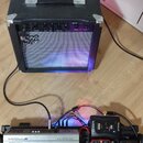 Černý kytarový kombozesilovač s nápisem SRV na předním mřížkovém krytu. Před ním leží stříbrný pedál s nápisy MODULE, TERRA NOVA a BASS PREAMP PEDAL.…