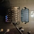 Detailní pohled na černý trup elektrické kytary s instalovaným tremolo mostem a snímačem. Na snímači je viditelný nápis VE. Povrch kytary vykazuje zn…