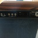 Záběr na zadní panel černého elektronického klavíru s dřevěným povrchem v horní části. Na panelu jsou umístěny konektory pro TREMOLO, SUSTAIN (FOOT S…