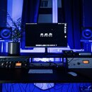 Fotografie zachycuje domácí studio s černým pracovním stolem a modrým osvětlením. Na stole stojí monitor s nápisem „s p h RECORDING“, dvě studiové mo…