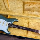 Elektrokytara s modrozeleným metallic povrchem a bílým pickguardem v otevřeném kufru s žlutou výplní. Na hlavě hmatníku je nápis Fender STRATOCASTER.…