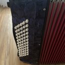 Detailní pohled na levou stranu akordeonu s tmavomodrým mramorovaným povrchem a červenými měchy. Na přední části je vidět sada bílých kulatých basový…
