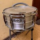 Stříbrný buben s kovovým obručováním a bílým blanou značky EVANS. Na korpusu je umístěn štítek s nápisy Pearl, SENSITONE a HERITAGE SERIES. Povrch bl…
