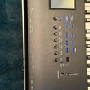 Detailní pohled na ovládací panel černé klávesové pracovní stanice. Na displeji je vidět nápis NAUTUS MUSIC WORKSTATION. Panel obsahuje několik otočn…