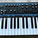 Černý syntezátor s klávesnicí a ovládacím panelem s modrými detaily. Na levé straně je bílý nápis novation a pod ním text Bass Station II analogue sy…