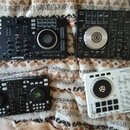 Fotografie zobrazuje čtyři DJ kontrolery položené na vzorité přikrývce. V horní levé části je černý kontroler s nápisy serato a Numark. V horní pravé…