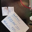 Fotografie zachycuje detail vnitřku elektronického zařízení s černým šasi a červeným kabelem. Na povrchu jsou přilepené tři bílé papírové štítky s ru…