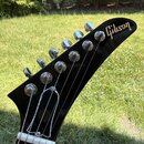 Detail hlavy elektrické kytary v černé barvě s chromovanými ladicími kolíky. Na vrchu je bílý nápis Gibson. Povrch lakovaný černou barvou vykazuje dr…