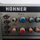 Detailní pohled na ovládací panel přístroje značky HOHNER v stříbrném provedení. Panel obsahuje čtyři vstupní konektory, jeden potenciometr s bílým k…