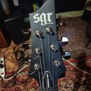Detail hlavy elektrické kytary v matně černé barvě. Na horní části je bílý nápis SGR by SCHECTER®. Na zadní straně krku je viditelný bílý štítek s ná…