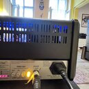 Zadní strana černého elektronického zařízení s ventilační mřížkou a modrým podsvícením uvnitř. Na kovovém panelu jsou nápisy TSC, POWER SOAK a SPEAKE…