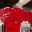 Záběr na zadní stranu červené hlavy kytary. Uprostřed je vyrazeno sériové číslo 81738501 a níže nápis MADE IN USA. Na pravém okraji hlavy je viditeln…