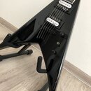 Černá elektrická kytara s ostrým, trojúhelníkovým tvaru těla umístěná na černém stojanu. Nástroj má dva snímače typu humbucker, na kterých je uveden…