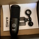 Černý kondenzátorový mikrofon s nápisem AKG P120 a logem značky. K mikrofonu patří černý popfiltr a šedý držák. Vše je umístěno v krabici na bílém pa…