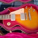 Elektrická kytara s tvarem singlecut a barevným přechodem od červené po žlutou leží v otevřeném kufru s růžovou výstelkou. Nástroj má dva humbuckery,…