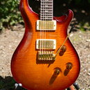 Elektrická kytara s červeně hnědým lakovaným povrchem z plamenového javoru. Nástroj má dva zlaté humbuckery, zlatý most s páčkou tremola a tři zlaté…