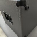 Detailní pohled na roh šedého reproduktorového boxu potaženého texturovaným materiálem. V horním rohu je přišroubován černý plastový ochranný roh a v…
