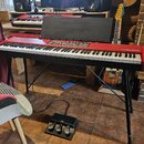 Červený digitální klavír s nápisem nord piano na černém stojanu. Nástroj má v horní části ovládací prvky a umístěný černý notový stojan. V popředí na…